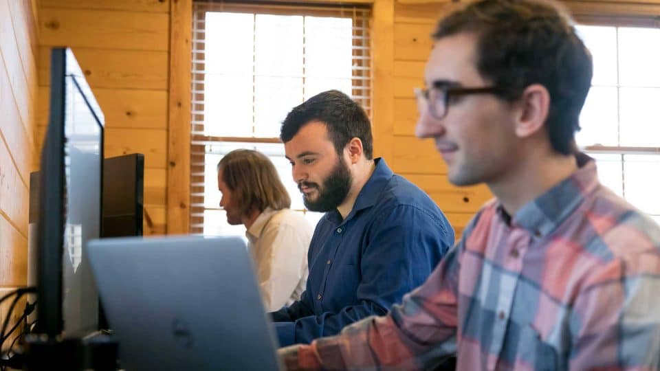 Three people working on laptops in a wooden office with windows.