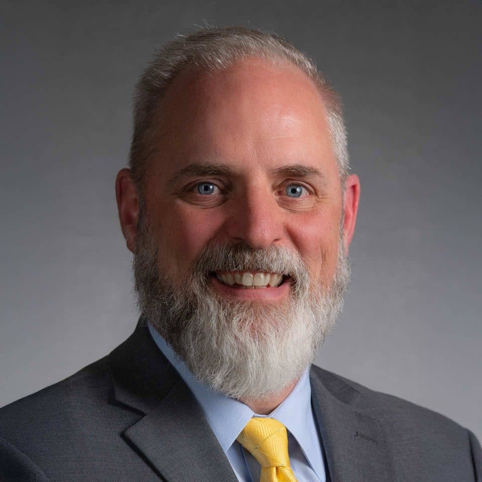 Smiling man with a gray beard in a suit and yellow tie against a gray background.